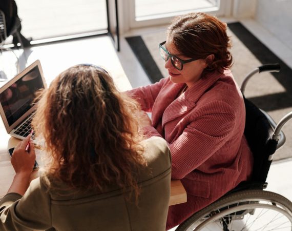 Dos mujeres, una de ellas en silla de ruedas, trabajando juntas en un portátil, representando la inclusión y accesibilidad en los servicios legales.